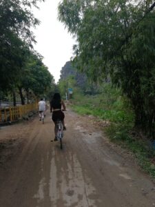 Cyclist on Cat Ba Island trail with Pelican Cruises boat docked, eco-friendly Ha Long Bay cruises