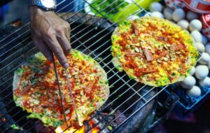A sizzling banh trang nuong being grilled over charcoal at the night market, topped with egg, scallions, and chili sauce, with a vendor’s hands in action.